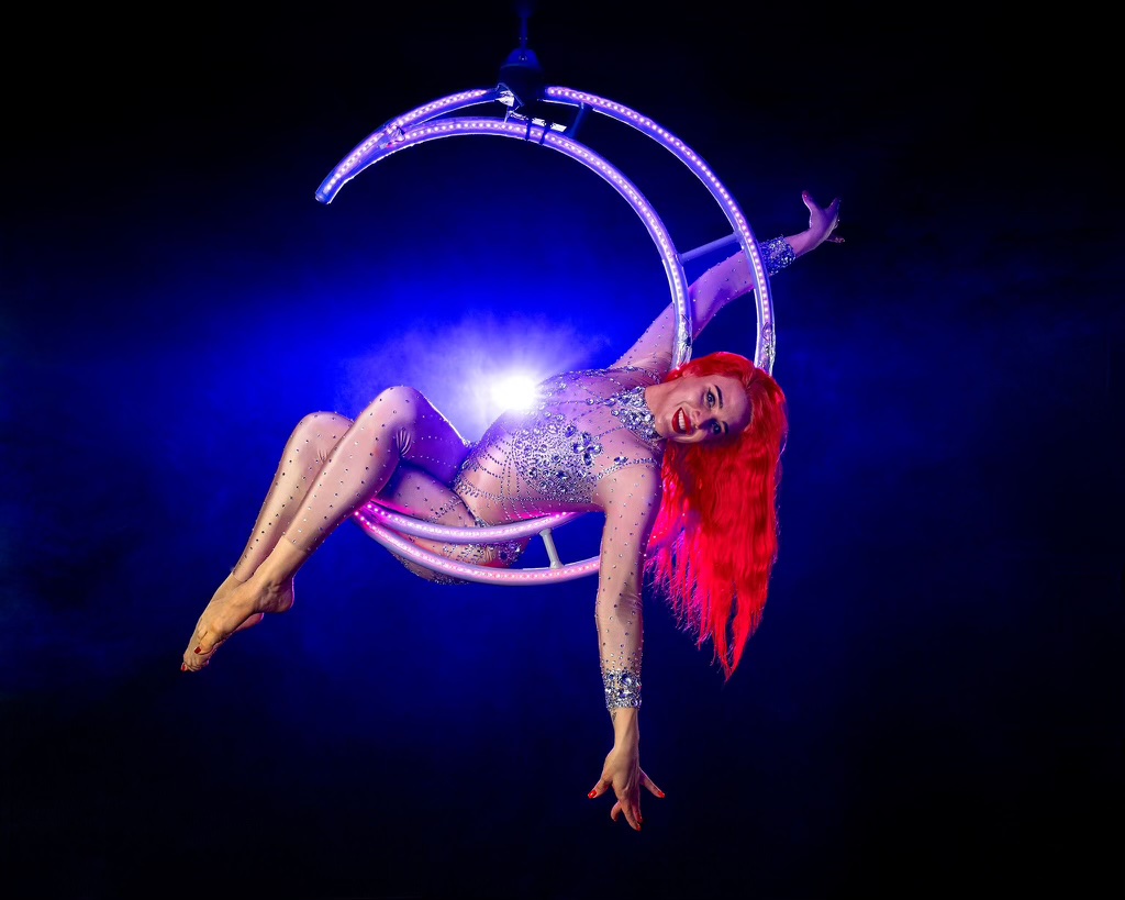 Red-haired aerialist reclining on a purple illuminated crescent moon apparatus against blue lighting